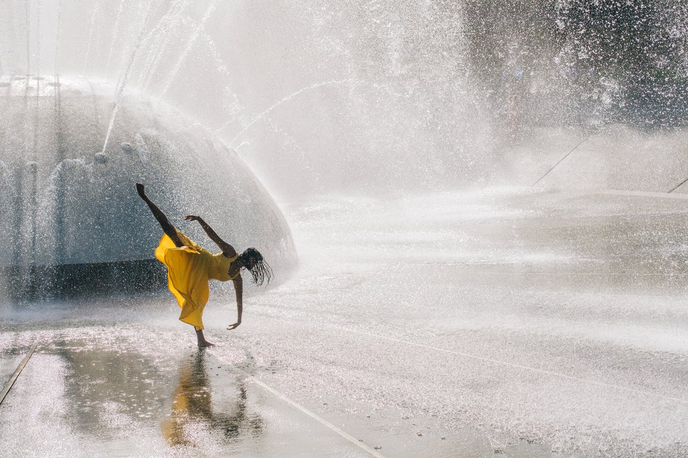 Woman barefoot in a yellow dress dances at a large outdoor water fountain in the USA on a sunny summer day.