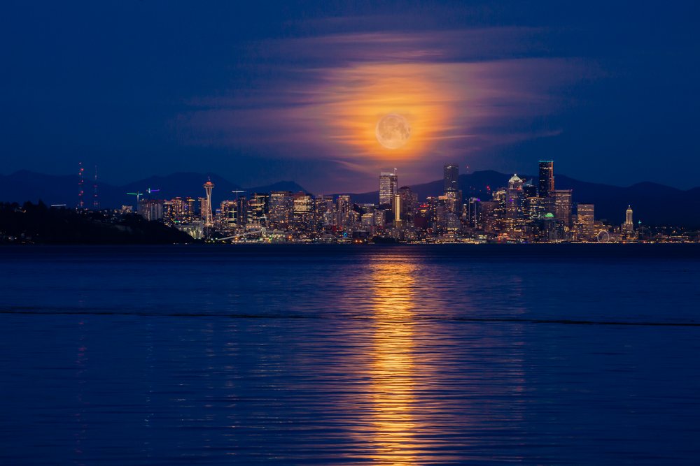Full moon illuminates Seattle skyline, reflecting on calm water at night with mountains and Space Needle visible, USA.