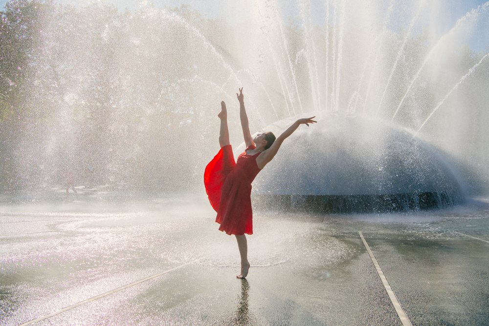 Woman in a red dress performs a graceful dance pose barefoot by a large outdoor water fountain with water spray and sunlight, USA.