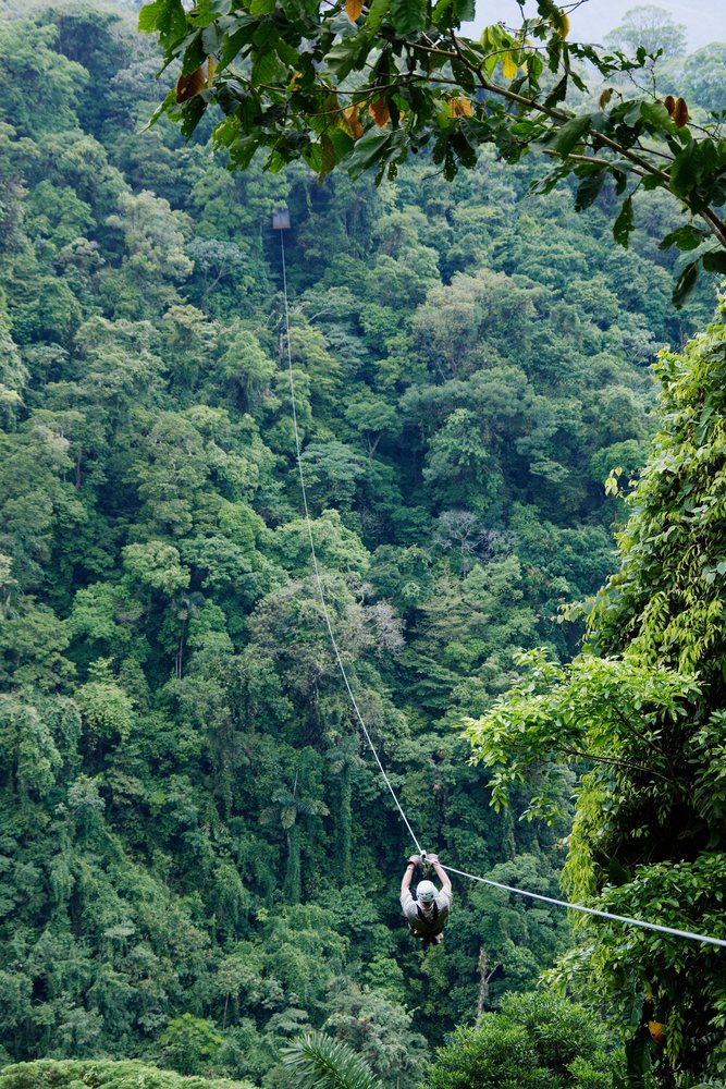 Person wearing a helmet descends on a zip line over a dense tropical forest canopy in Costa Rica.