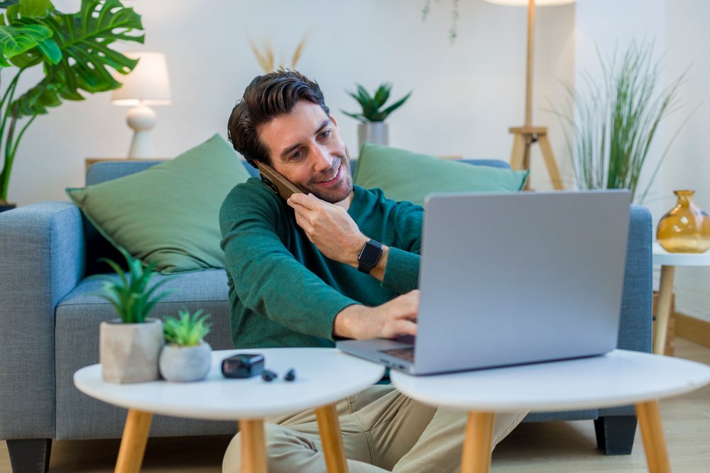 Man sitting on floor in front of couch using laptop and talking on smartphone in a modern living room with indoor plants, technology, young adult, home.