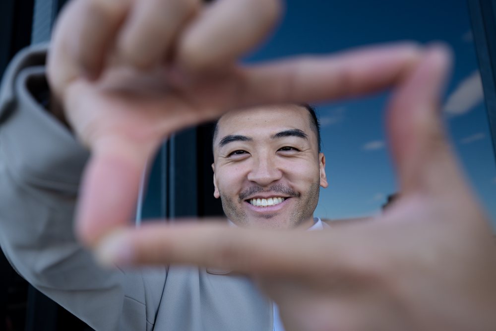 Man smiling and framing his face with hands outdoors against blue sky and window reflection, Madrid, Spain.