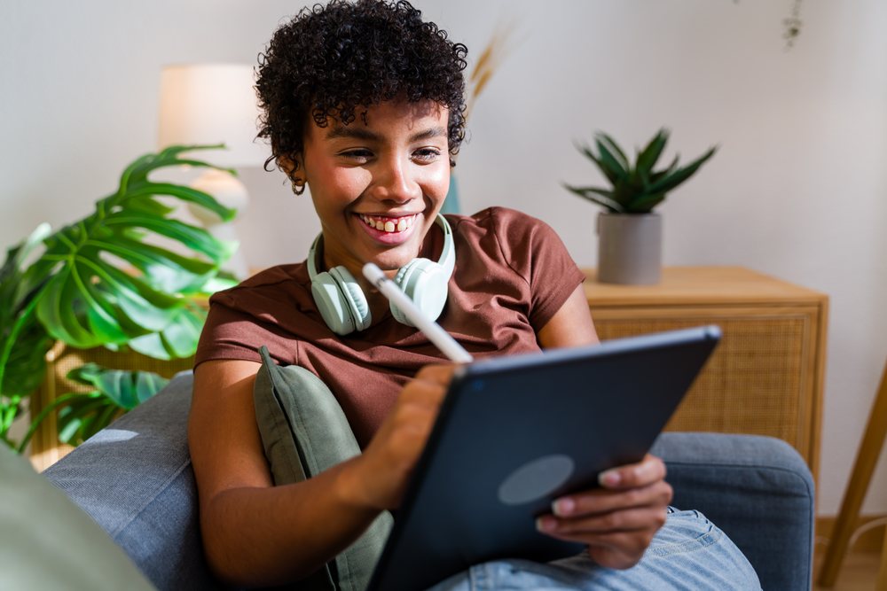 Woman with curly hair smiling while using a tablet with stylus pen on a sofa near indoor plants in a living room, Spain.