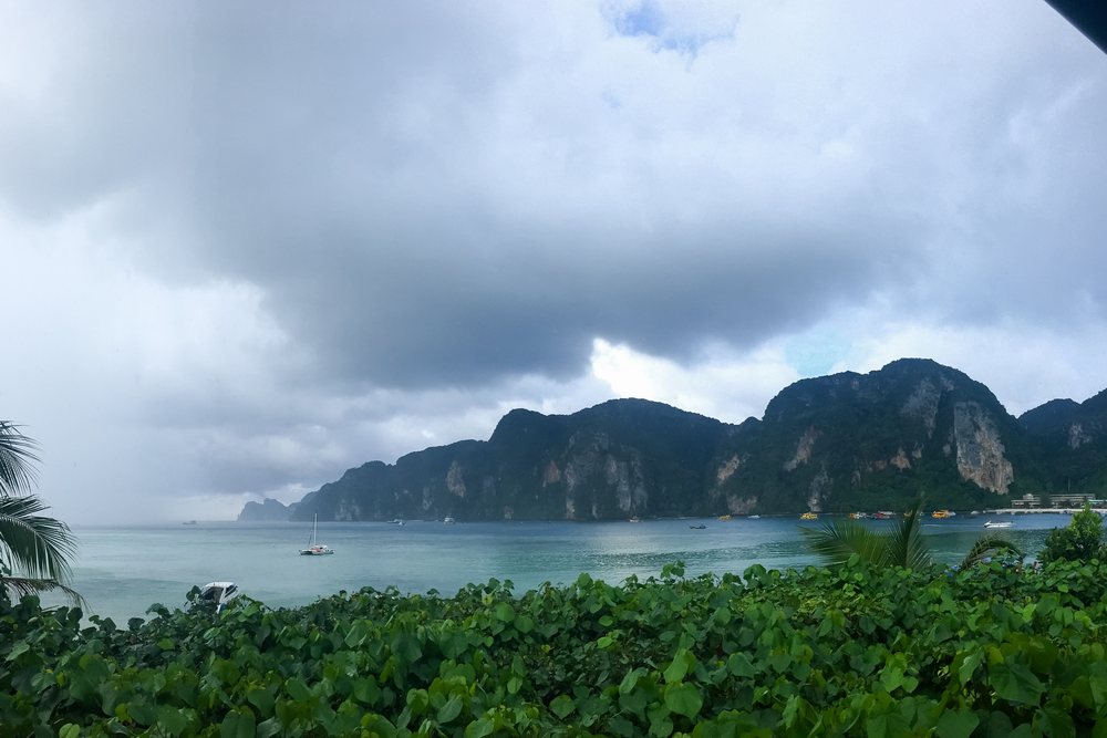 Calm tropical coastline in the Philippines with lush vegetation, sailboats, speedboats, and island cliffs under an overcast sky with rain clouds.
