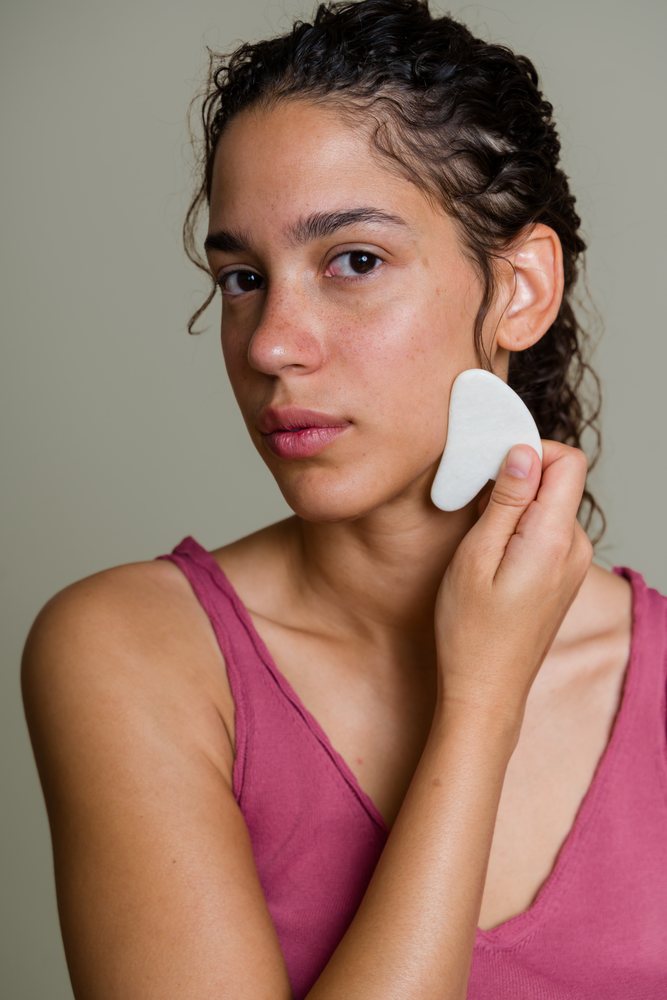 Woman performs a facial massage on her face using a white gua sha tool against a neutral background, beauty shoot, Spain.