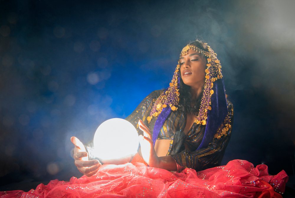 A woman dressed in mystical attire holds a glowing crystal ball in a dark, smoky indoor setting in the, fortune teller, spirituality, USA.