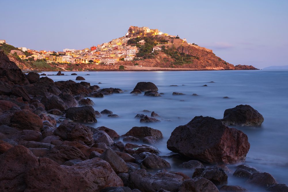Hilltop castle overlooking Castelsardo seaside town at Dawn with rocky shore and illuminated buildings along Sardinia's Mediterranean coast, Travel Images, Italy.