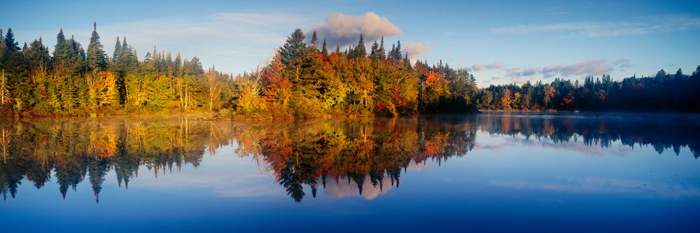 Colorful autumn trees with red, orange, and yellow leaves reflect in a calm lake under a partly cloudy sky with light mist over the water.