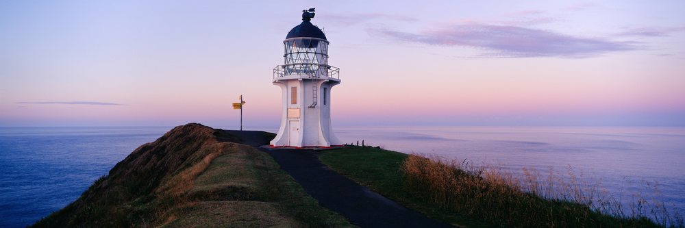 White lighthouse on a coastal cliff at sunset with ocean view, a grassy path, and a signpost under a pink and purple twilight sky.