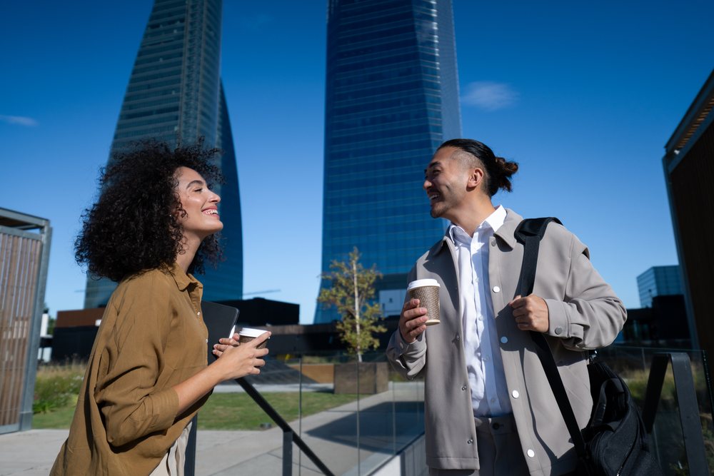 Two young adults smiling and talking outdoors near modern glass skyscrapers, each holding a disposable coffee cup on a sunny day, Madrid, Spain.