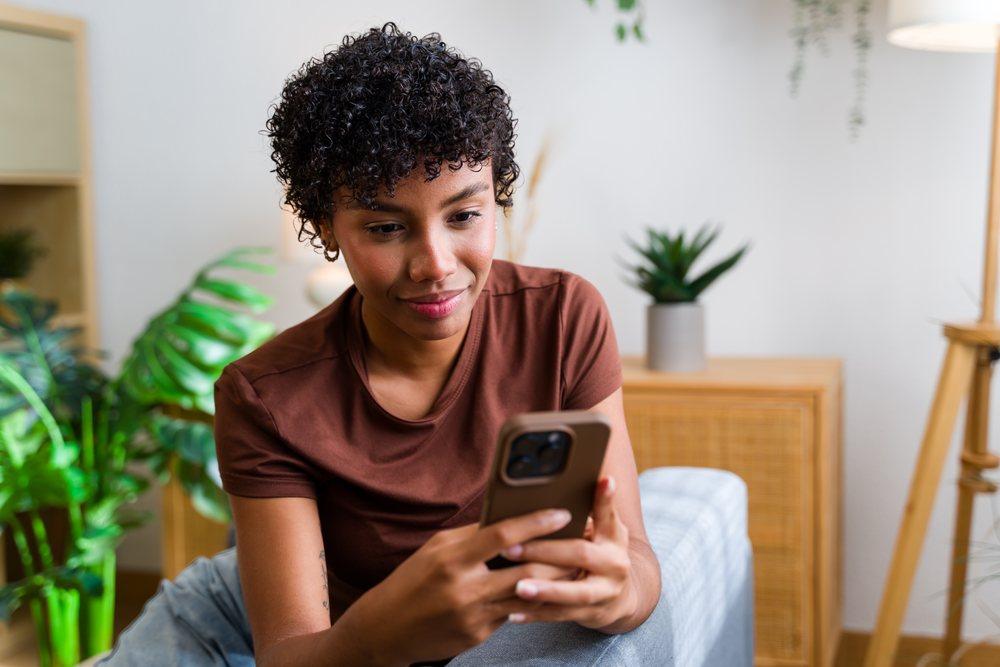 Young woman with curly black hair sits indoors looking at her smartphone screen, with a houseplant and wooden furniture in the background, Spain.