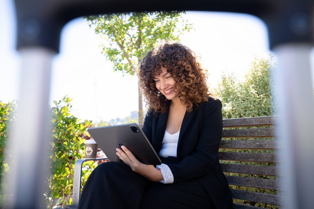 Smiling woman with curly hair uses a tablet while sitting on a wooden bench outdoors in sunlight near green plants and a coffee cup, Spain.