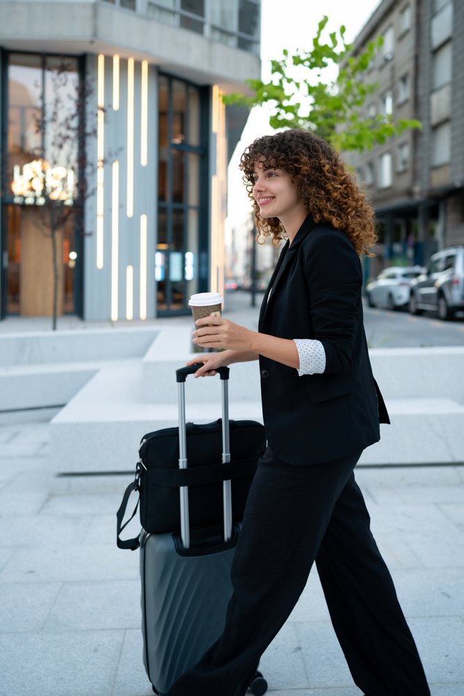 Business woman with curly hair walks on an urban sidewalk in Madrid pulling rolling luggage and holding a disposable coffee cup.