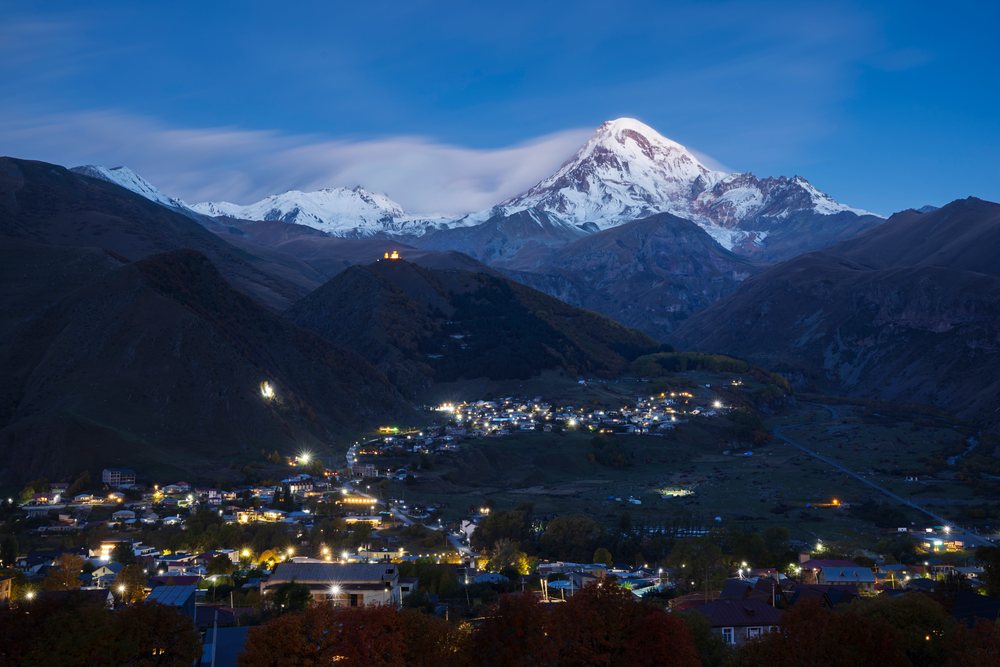 Mt Kazbegi rises over Stepantsminda village at Dawn with snow-capped peaks and an illuminated hilltop church, Georgia.