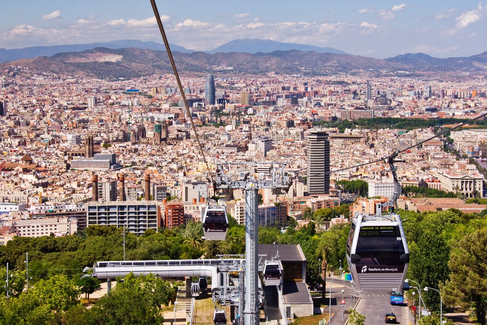 Cable car cabins of Telefèric de Montjuïc travel over urban greenery toward Barcelona cityscape with mountains beyond, Spain.
