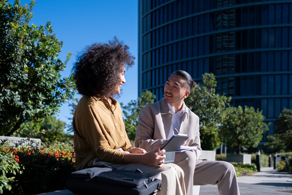 Two business professionals engage in conversation while sitting on a bench in an urban park in Madrid during daylight, Spain.