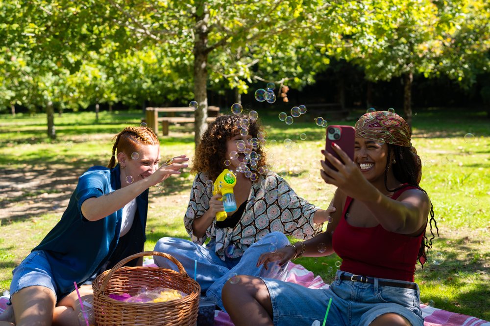 Three women sit on a picnic blanket outdoors, playing with bubbles and taking a selfie, Spain.