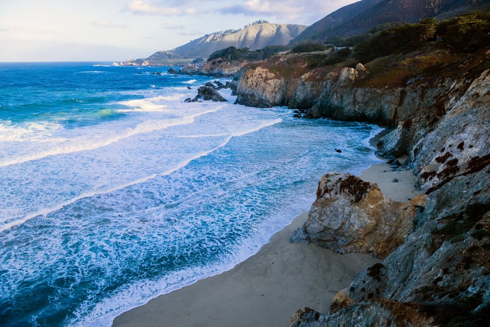 Waves crash along the rocky coastline and sandy beach with cliffs and coastal vegetation, California, USA.