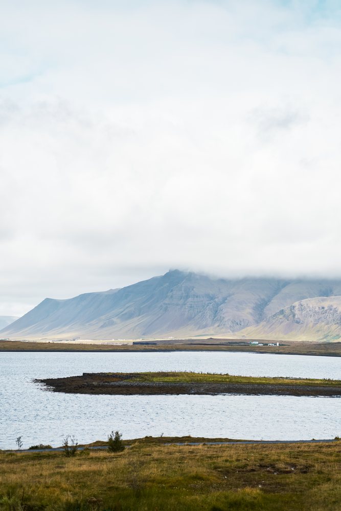 A lake with a small island and grassy field in front of a mountain partially covered by clouds, Iceland.