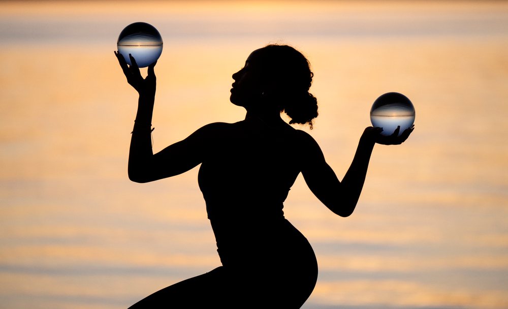 Woman balanced in yoga pose holding two glass balls at sunset with water background, USA.