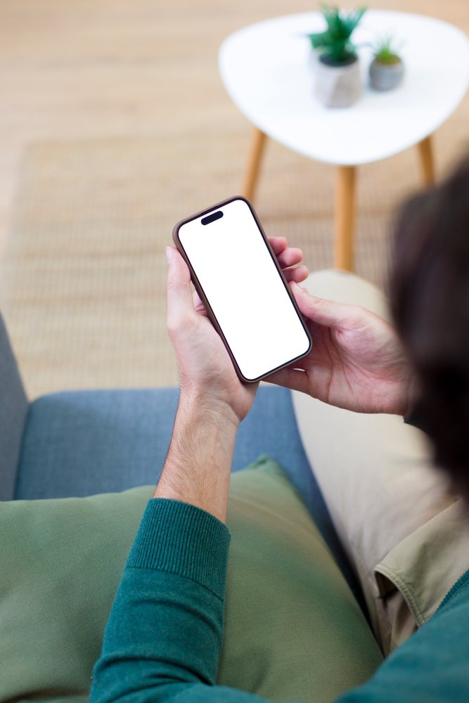 Person seated on a blue couch holding a smartphone with a blank screen near a white table topped with two potted plants in a home living room, technology, young adult.