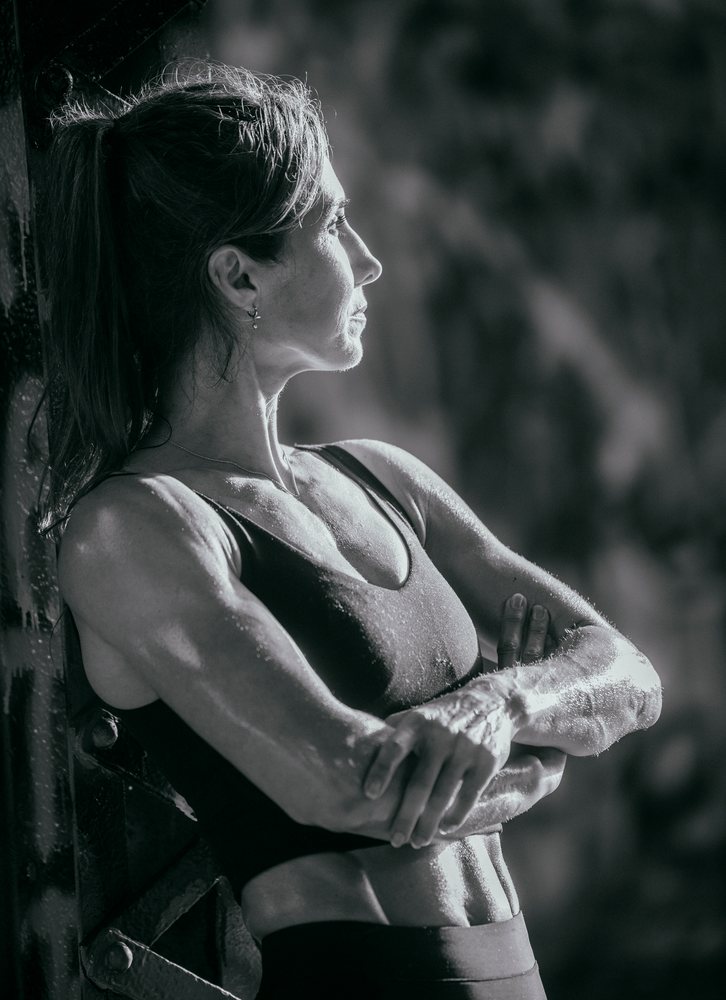 Muscular woman with hair tied back stands sweating in profile during outdoor calisthenics workout, USA.