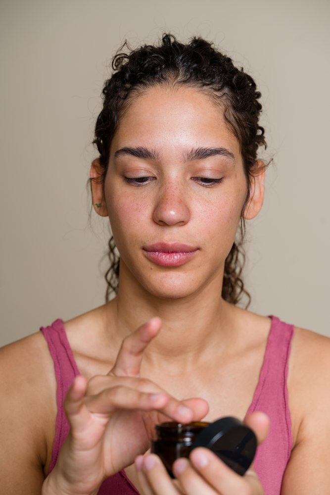 Woman with curly hair applying cream from a brown jar during a skincare beauty shoot, Spain.