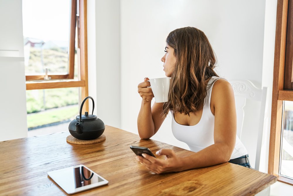 A white woman enjoys coffee and checks her phone by a wooden table
