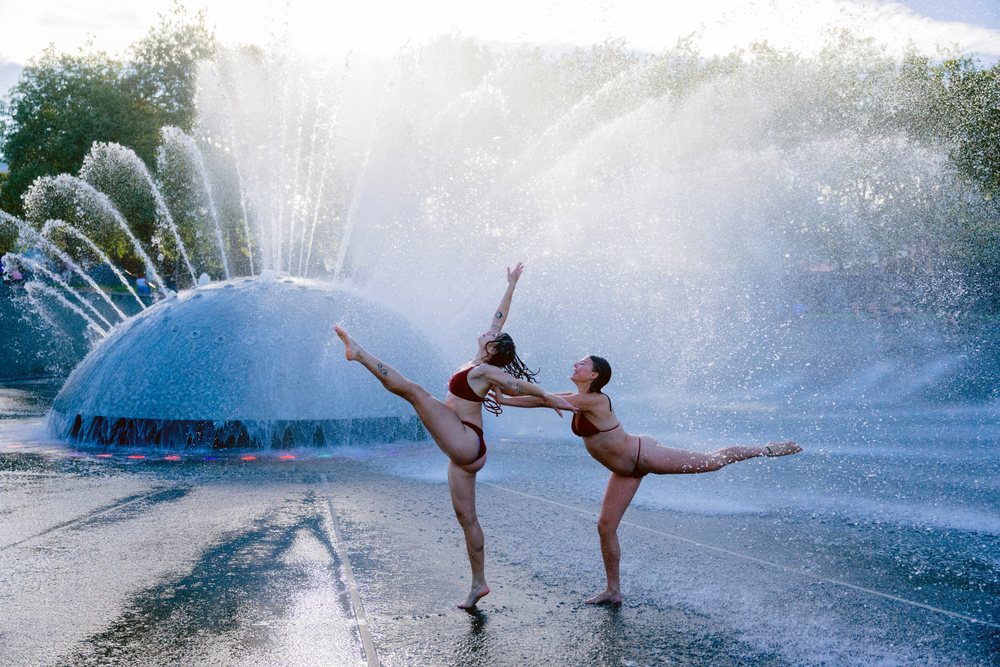 Two women wearing bikinis performing acrobatic yoga poses together in front of a large water fountain with mist and spray in an outdoor setting, partner, acrobatics, USA.