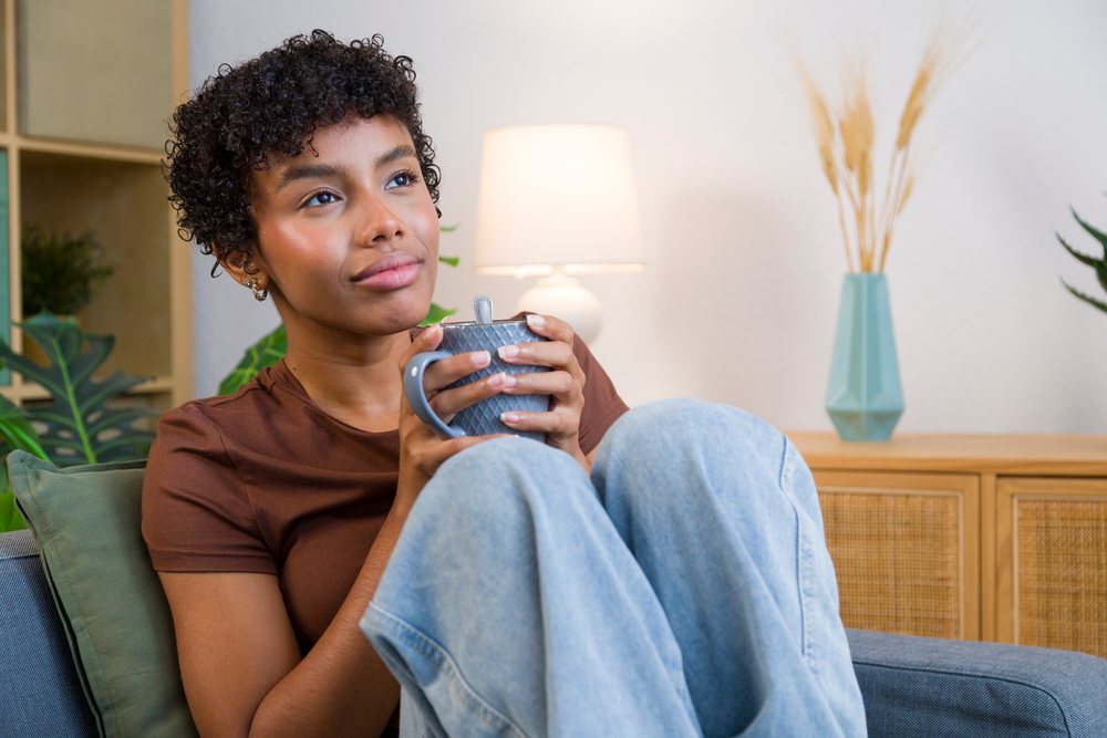 Young woman sitting on a sofa holding a coffee mug in a cozy home interior with decor and plants.