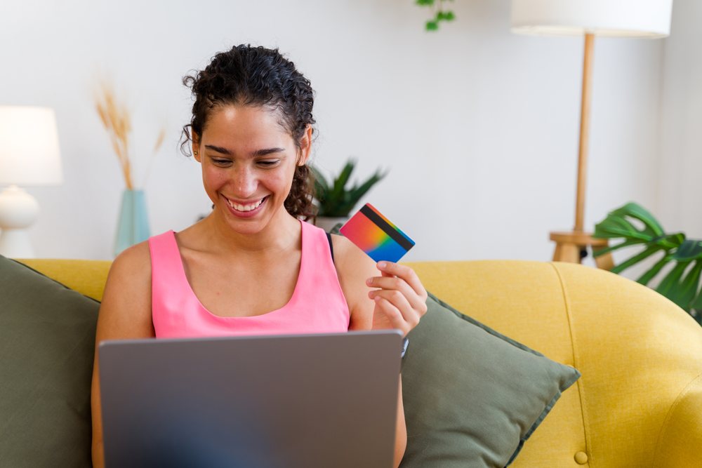 A woman holding a credit card and using a laptop for online shopping while sitting on a yellow sofa in a modern home interior, Spain.
