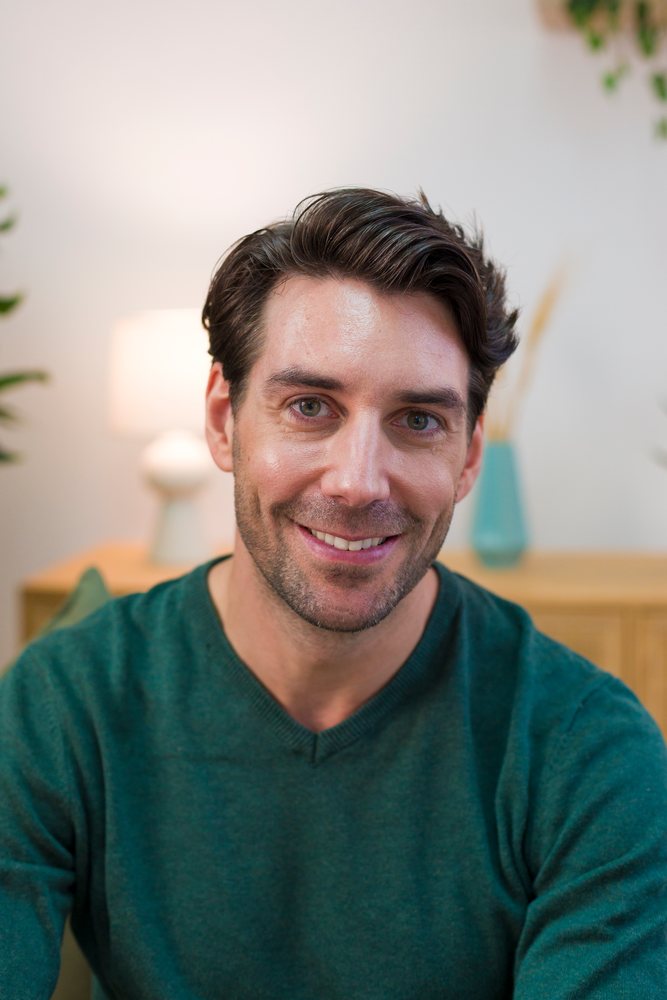 Young man smiling indoors in casual home setting wearing a green sweater, Technology, young adult.