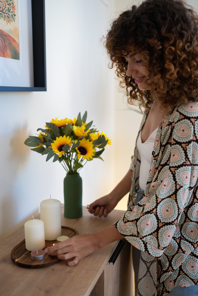 Woman with curly hair lights a tea light candle on a wooden tray with yellow sunflowers in a green vase beside her indoors, home, girl, Spain.