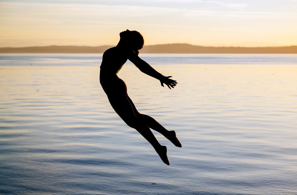 Woman performing a yoga-like jump by a lake at sunset, USA.