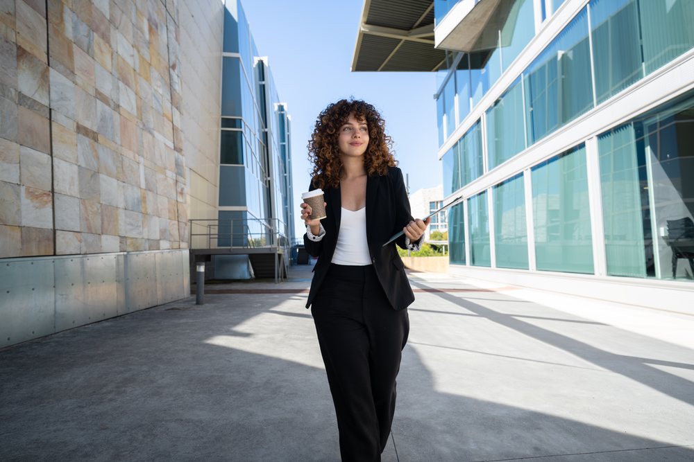 Businesswoman walking outdoors holding a coffee cup and folder between modern office buildings, traveling executive, Spain.