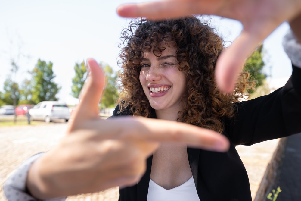 Woman with curly hair winking and making a framing gesture with her hands outdoors, business professional, business traveler, Spain.