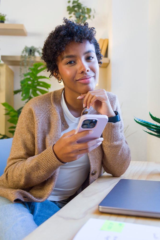 Young woman sitting at a desk using her smartphone with indoor plants and a laptop nearby in a home setting.