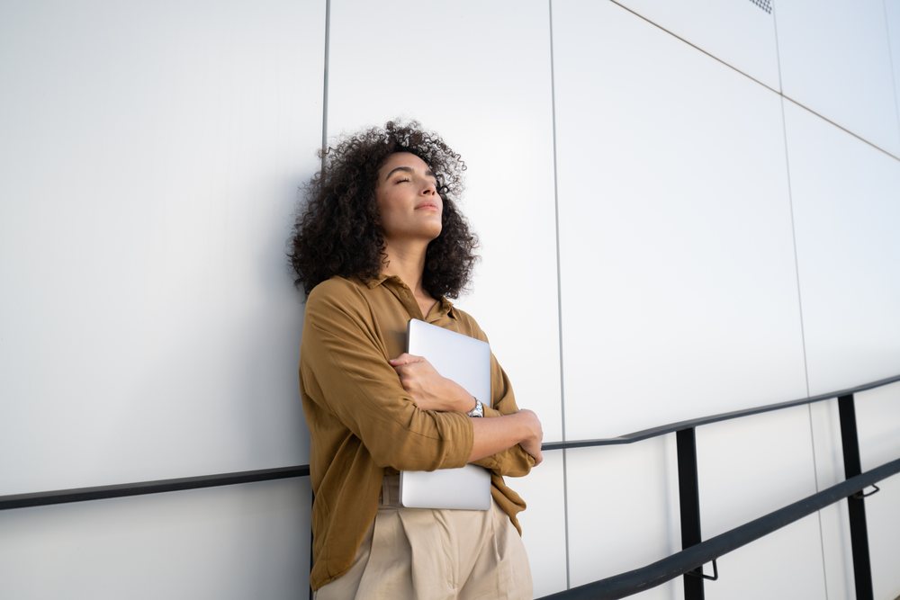 Woman with closed eyes holds a laptop while leaning against a modern building wall outdoors, Madrid, Spain.