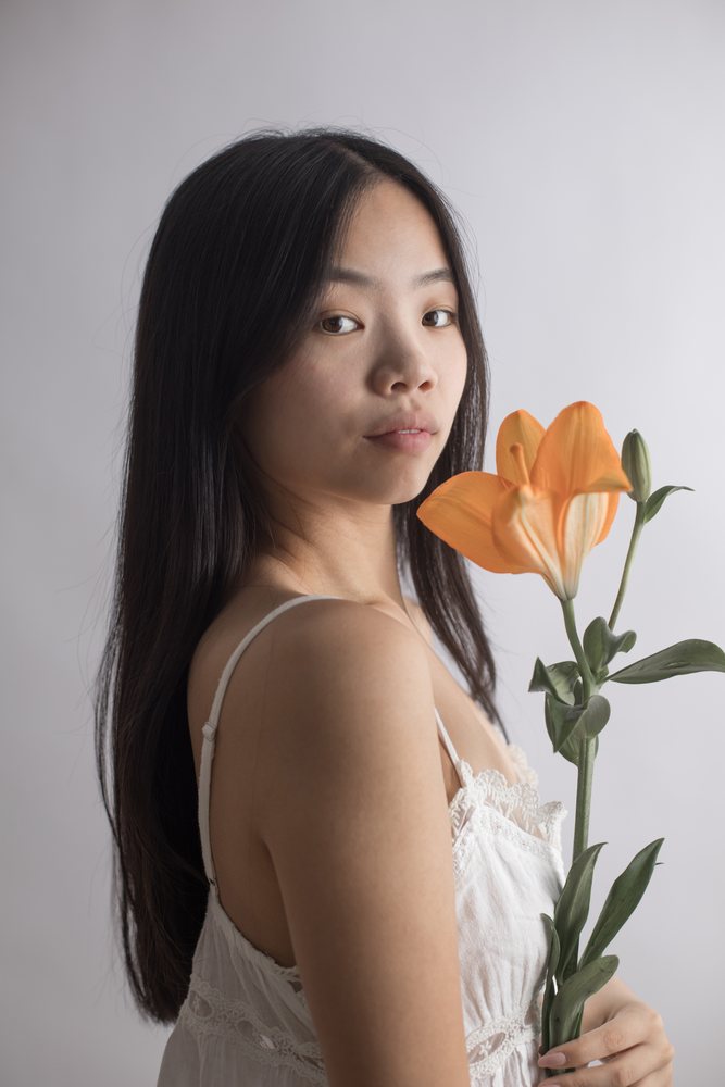 Studio portrait of a woman with long dark hair holding an orange lily flower against a white background, Art, emotional.