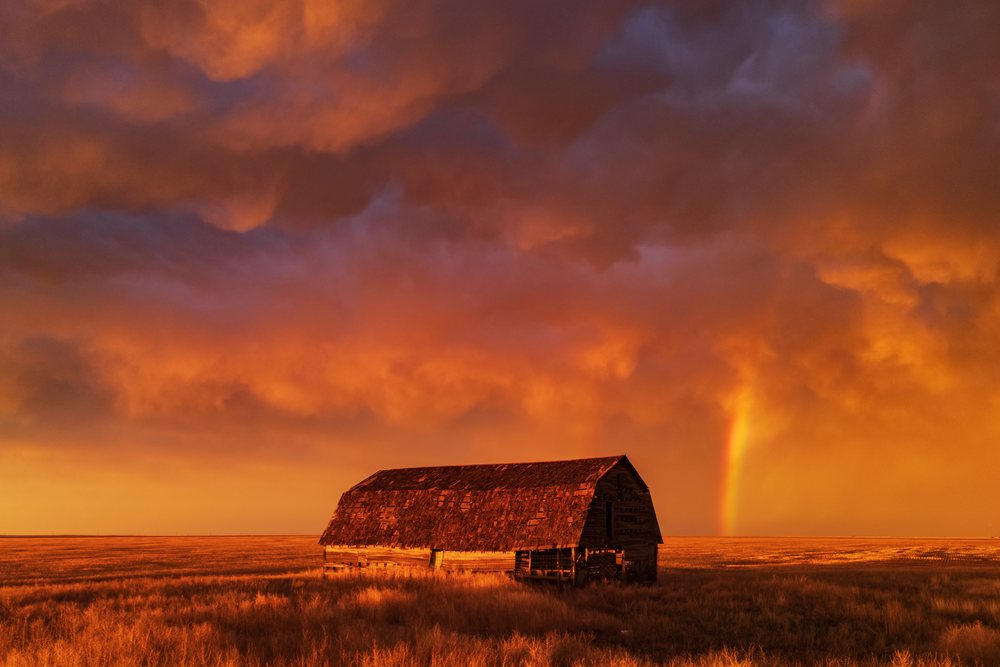 Old wooden barn stands in a dry grassy field under orange and purple storm clouds at Sunset with a visible Rainbow segment, Storm Chasing, Weather, Environment, Eva, Oklahoma, USA.