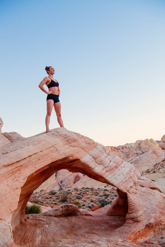 Woman barefoot in a yoga pose standing confidently on a natural sandstone rock arch in a desert landscape at sunset, USA.
