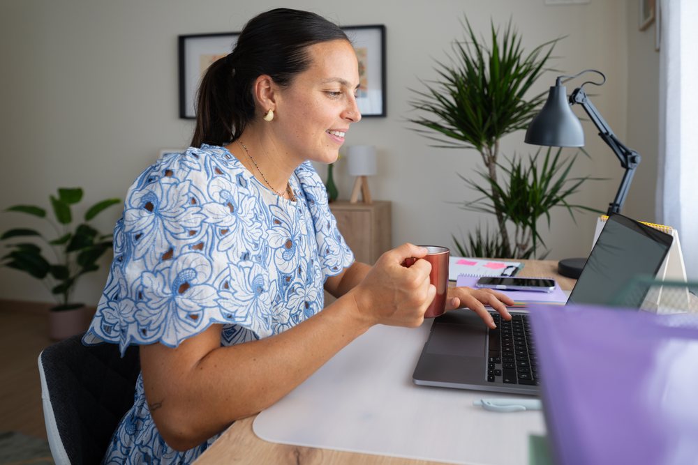 Woman drinking coffee and working on a laptop at a home office desk near a window, telecommuting, Spain.