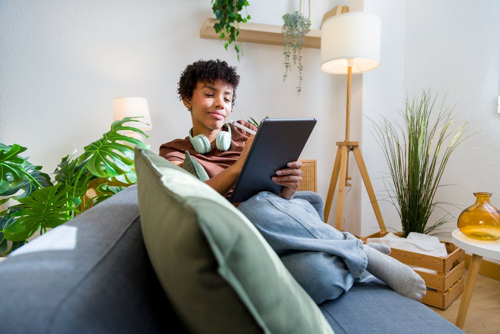 Young woman wearing earphones sits on a gray sofa holding and writing on a tablet in a modern living room with houseplants and decorative objects.