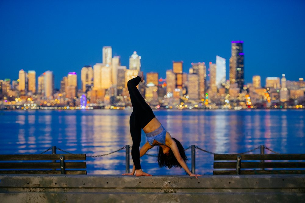 Woman performing yoga backbend on a waterfront bench with the Seattle skyline visible at dusk, WA, USA.