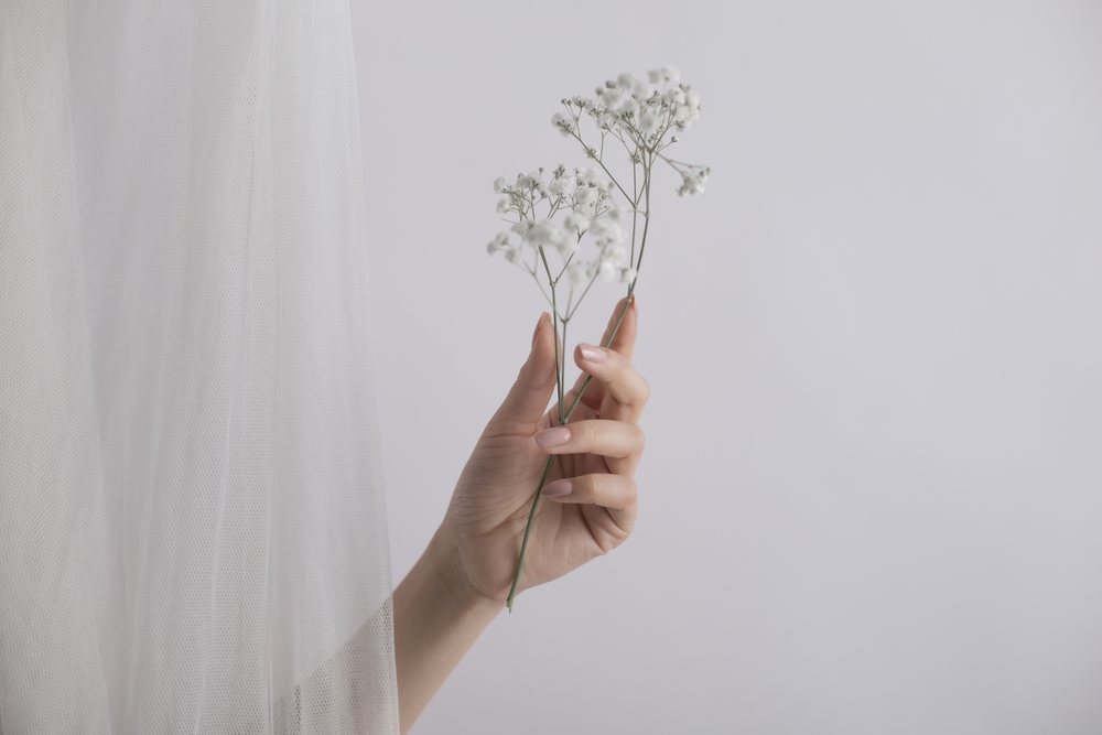 Human hand delicate white baby's breath flowers beside white tulle veil fabric against a light neutral background, Art, portrait, emotional.