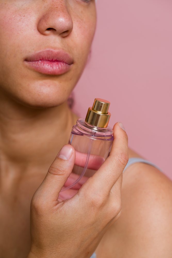 Woman holding a clear perfume bottle with a golden spray top near her face against a pink background during a beauty shoot, Spain.