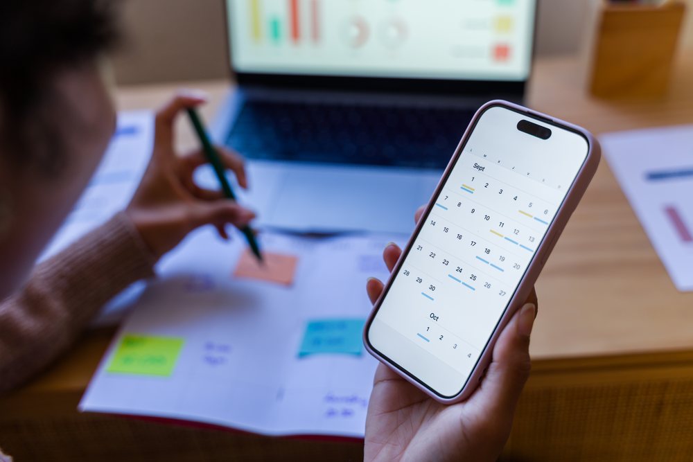 Person holding smartphone showing calendar app for September and October, writing with pen on planning notes beside a laptop on a desk, Spain.