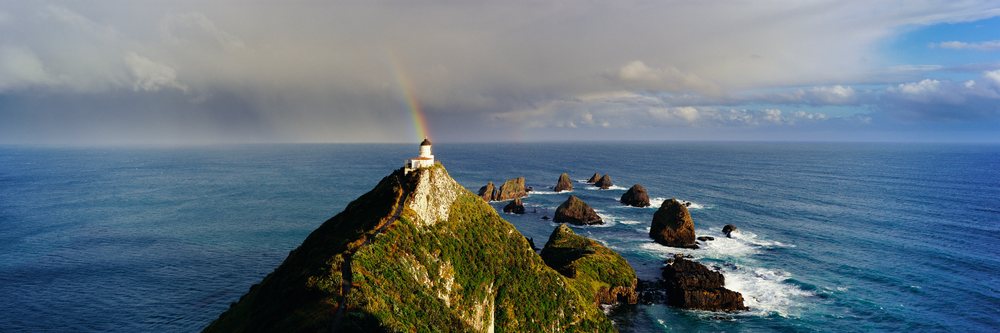 Lighthouse on a cliff at a rocky ocean coastline with a rainbow arching above under a partly cloudy sky.