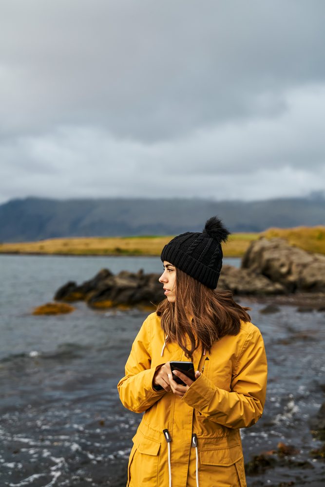 A white woman in a yellow jacket uses a smartphone by the shore