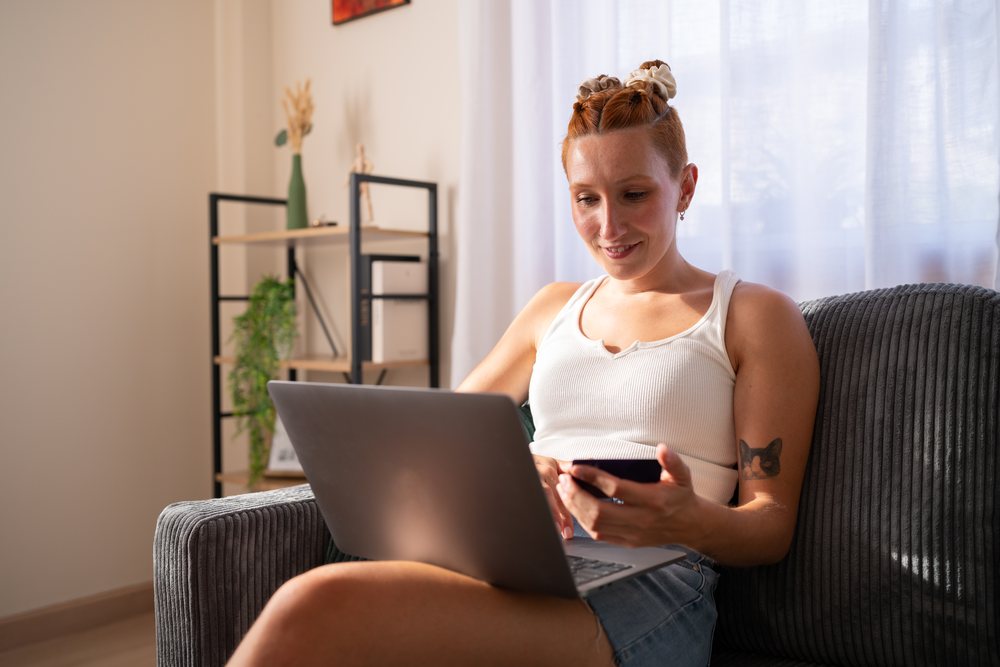A woman using a laptop and holding a payment card while sitting on a couch at home, moving, Spain.